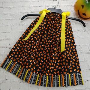 K&ouml;nnte beinhalten: Ein schwarzes und oranges Kleid mit einem Candy-Corn-Druck und gelben Schleifenb&auml;ndern. Das Kleid hat einen gestreiften Saum mit schwarzen, orangen, gelben und grauen Streifen.