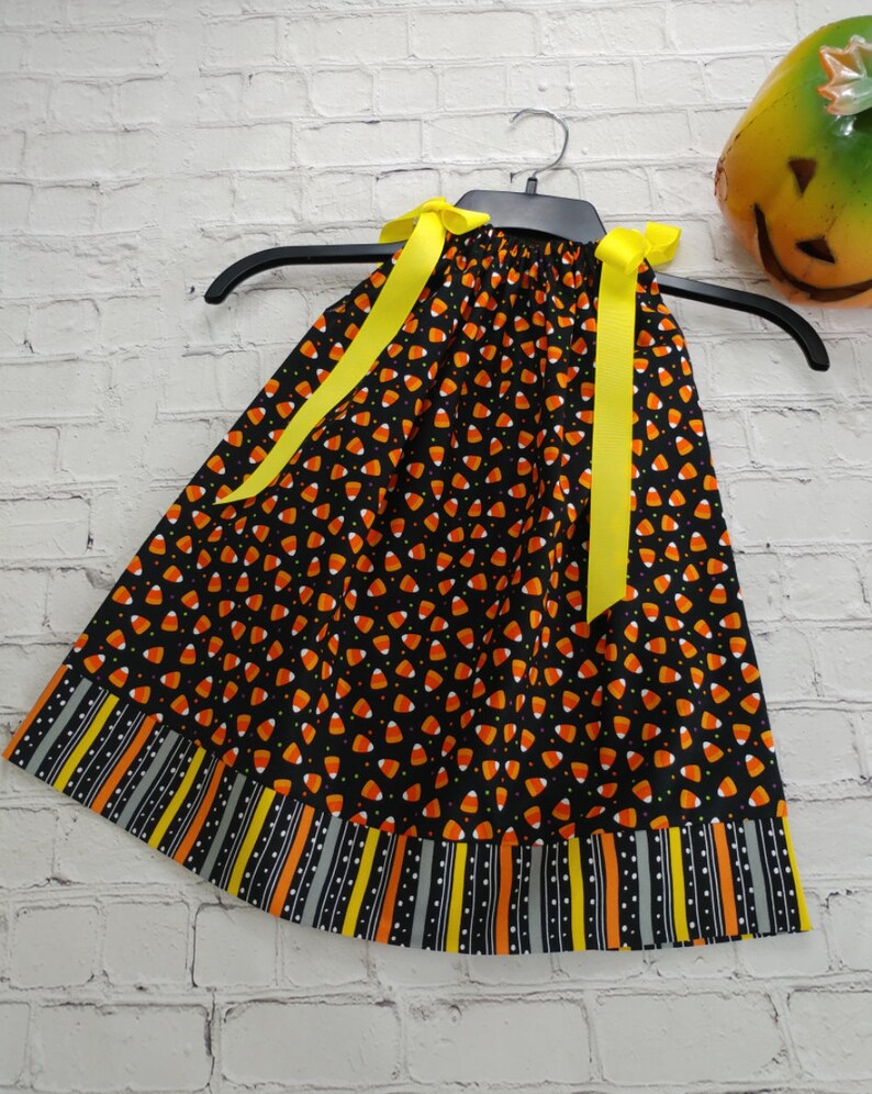 K&ouml;nnte beinhalten: Ein schwarzes und oranges Kleid mit einem Candy-Corn-Druck und gelben Schleifenb&auml;ndern. Das Kleid hat einen gestreiften Saum mit schwarzen, orangen, gelben und grauen Streifen.