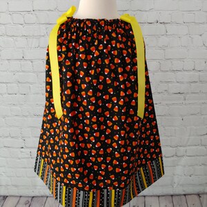 K&ouml;nnte beinhalten: Ein schwarzes und oranges Kleid mit einem Maisbonbon-Druck und gelben Schleifenb&auml;ndern. Das Kleid hat einen schwarzen, orangen und wei&szlig;en Streifenbesatz am Saum.