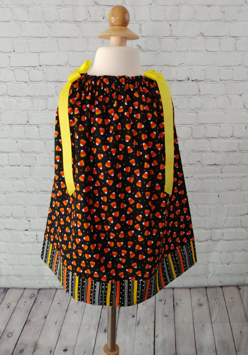 K&ouml;nnte beinhalten: Ein schwarzes und oranges Kleid mit einem Maisbonbon-Druck und gelben Schleifenb&auml;ndern. Das Kleid hat einen schwarzen, orangen und wei&szlig;en Streifenbesatz am Saum.