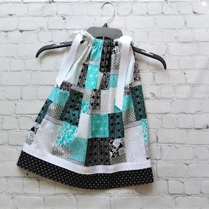 May include: A patchwork dress with a white ribbon and black polka dot trim. The dress is made of various fabrics in shades of teal, black, white, and gray.