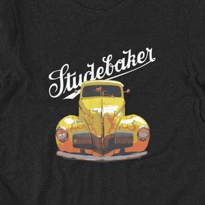 Könnte beinhalten: Dunkelgraues T-Shirt mit einem Vintage-Studebaker-Auto-Motiv in Gelb und Orange. Das Wort "Studebaker" steht in weißer Schrift über dem Auto. Das Shirt besteht aus einem weichen Material.