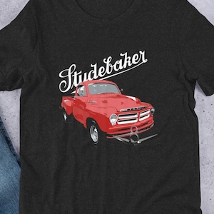 Könnte beinhalten: Ein schwarzes T-Shirt mit einem roten Studebaker-Pickup-Truck-Motiv und dem Text "Studebaker" in weißer Kursivschrift.