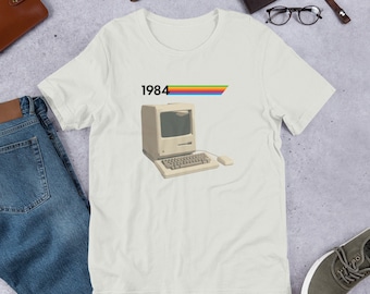 Puede incluir: Una camiseta blanca con un gráfico de computadora vintage y el texto "1984" encima. La computadora es beige con una franja de arcoíris encima.