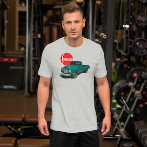 May include: A light grey short-sleeved t-shirt featuring a graphic of a vintage green pickup truck positioned below a red circular logo containing the white text STUDEBAKER CARS & TRUCKS. The shirt has a crew neckline and a standard fit.