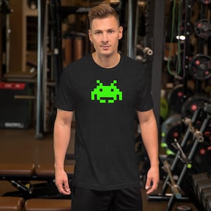May include: Dark gray t-shirt featuring a pixelated green alien graphic. The short-sleeved tee is made of a soft material and is suitable for casual wear. The design is a nod to retro video games.