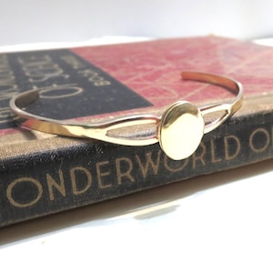 May include: A gold-toned cuff bracelet with a circular centerpiece. The bracelet is resting on a vintage book with the title "Wonderworld of Science" visible. The bracelet has a simple, elegant design.