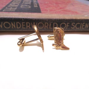May include: A pair of gold-toned cufflinks featuring a cowboy boot design. The boots have a detailed pattern and a spur on the heel.