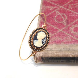 Könnte beinhalten: Ein goldfarbenes Armreif-Armband mit einem Cameo-Anhänger. Der Anhänger hat einen schwarzen Hintergrund mit einem weißen Profil einer Frau. Der Cameo ist von einem goldfarbenen Perlenrand und einem schwarzen Rand umgeben. Das Armband liegt auf einem alten Buch.