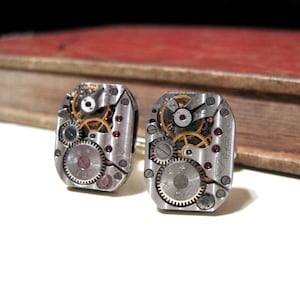 May include: A pair of silver cufflinks with exposed clockwork gears and a red jewel accent. The cufflinks are made from recycled watch parts.
