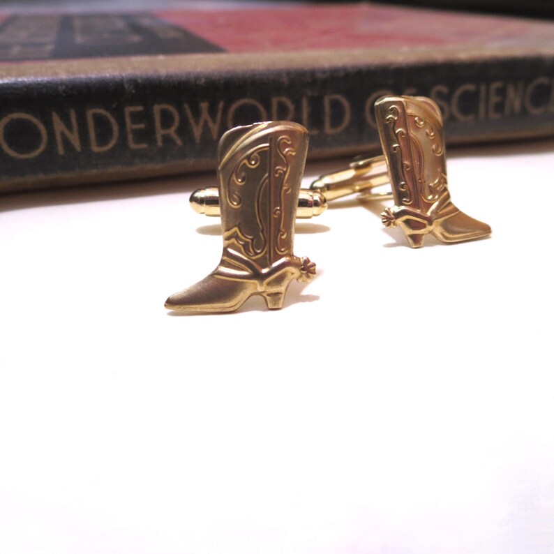 May include: A pair of gold-tone cufflinks featuring a cowboy boot design. The boots have a detailed design and a spur on the heel.