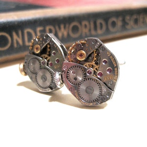 May include: A pair of silver cufflinks with exposed watch gears and ruby-colored accents. The cufflinks are made from vintage watch parts and have a unique steampunk aesthetic.