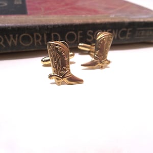 May include: A pair of gold-tone cufflinks shaped like cowboy boots. The boots have a detailed design and a spur on the heel.