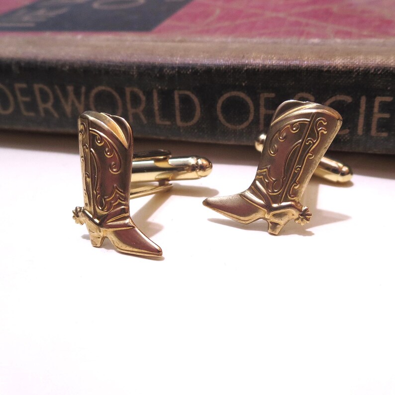 May include: A pair of gold-toned cufflinks shaped like cowboy boots. The boots have a detailed design and a spur on the heel.