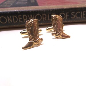 May include: A pair of gold-tone cufflinks featuring a detailed design of cowboy boots with a spur on the heel.
