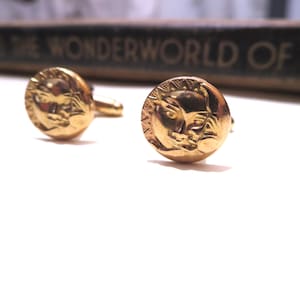 May include: A pair of gold-colored cufflinks. Each cufflink features a detailed relief of a face with a sunburst design around the edge. The cufflinks are round and have a classic, elegant design. The background includes a book with the title "THE WONDERWORLD OF".