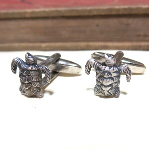 May include: A pair of silver cufflinks featuring a detailed turtle design. The turtles have textured shells and outstretched arms. The cufflinks have a classic, polished finish, perfect for adding a touch of personality to any formal attire.