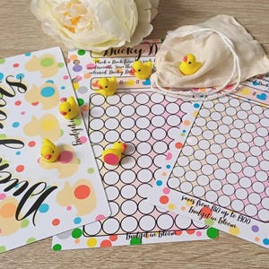 May include: Three rectangular paper savings trackers with colourful duck and polka dot patterns. The text reads Ducky Dots, Budget in Bloom, and Saves from £0 up to £100. Several small yellow rubber ducks are placed on the sheets and in a small cloth bag.