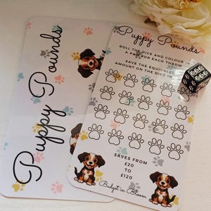 May include: Two white rectangular savings tracker cards titled Puppy Pounds with colourful paw print patterns and illustrations of puppies. One card features a grid of paw outlines for colouring, text instructions, and a black six-sided die with white paw pips.