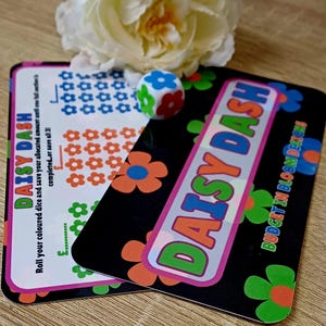 May include: A Daisy Dash savings challenge card set featuring a black card with orange and green flower graphics and the text Daisy Dash and Budget in Bloom Designs, plus a white card with rows of blue, orange, and green flower icons and a die.