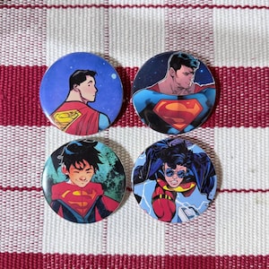 May include: Four circular pin-back buttons featuring comic book illustrations of Superman. The top left shows a profile view, the top right a chest-up portrait, the bottom left a smiling youth, and the bottom right an action pose with round glasses.