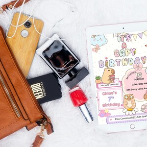 May include: A flat lay featuring a brown leather purse, a wooden-backed phone, a black perfume bottle, a black compact, and a red nail polish bottle. A tablet displays a birthday invitation with capybara illustrations and the text "It's a Capy Birthday!"