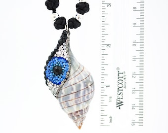 May include: A seashell pendant necklace with a black cord. The pendant is a natural shell, embellished with blue, black, and clear crystals in an eye design. The necklace measures approximately 3 inches in length.