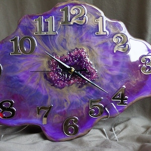 Puede incluir: Un reloj de resina morado y dorado con números plateados y una aguja negra. La esfera del reloj tiene forma de geoda con un patrón de remolino morado y dorado.