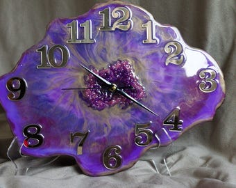 May include: A purple and gold resin clock with silver numbers and a black clock hand. The clock face is shaped like a geode with a purple and gold swirl pattern.