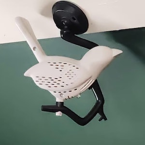 Op de afbeelding: Een witte vogelvormige wandlamp met een zwarte montagearm en voet. De vogel heeft een reeks kleine gaatjes op zijn lichaam. De lamp is gemonteerd op een muur met een groen en wit kleurenschema.