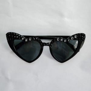 May include: Black heart-shaped sunglasses with rhinestones and the words "PRO CHOICE" spelled out in white letters.