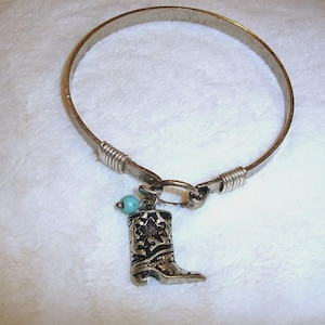 May include: Silver-toned bangle bracelet with a cowboy boot charm. The charm is detailed with decorative patterns and a small turquoise bead. The bracelet is made of metal and has a simple clasp.