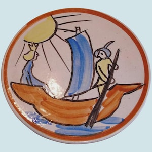 May include: A round ceramic plate featuring a hand-painted illustration of a sailboat on water with a yellow sun in the sky. The design includes two figures on the boat, rendered in blue, yellow, and brown outlines against a cream background.