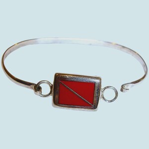 May include: Silver-toned bracelet featuring a rectangular red charm with a diagonal silver line. The bracelet has a simple, open design with a clasp. The charm is a vibrant red color, and the bracelet is made of a shiny metal.