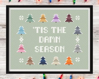 May include: Framed cross-stitch art with a light green background. The design features colorful Christmas trees and the text "'Tis the Damn Season" in white. The frame is black, and the background is decorated with Christmas lights.