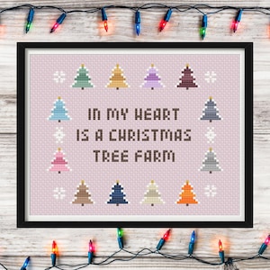 May include: Framed cross-stitch art with the text "IN MY HEART IS A CHRISTMAS TREE FARM" surrounded by colorful Christmas trees and snowflakes on a pink background. The frame is black, and the background is a light wood. Christmas lights are strung around the frame.