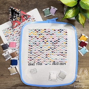 May include: A cross-stitch project in a blue hoop features a colorful heart pattern with the text "ERAS TOUR 2023-2024." The image includes spools of thread in various colors, a patterned fabric, and a small pin that says "It's me, hi, I'm the problem, it's me."