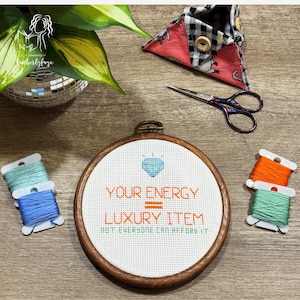 Puede incluir: Un bastidor de punto de cruz con el texto "YOUR ENERGY = LUXURY ITEM, NOT EVERYONE CAN AFFORD IT" en naranja. El diseño incluye un diamante azul. La imagen también muestra carretes de hilo, tijeras y un portaagujas de tela.