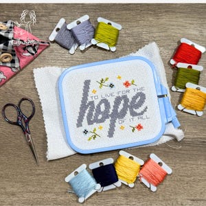 May include: A cross-stitch project with the words "hope" and "to live for the hope of it all" in gray thread. The project is in a blue hoop, surrounded by spools of colorful embroidery floss, scissors, and fabric.