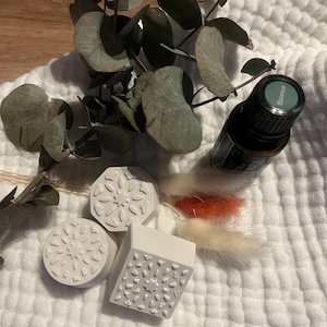 Könnte beinhalten: Ein Stillleben mit Wellness-Produkten. Eine dunkelbraune Flasche ätherischen Öls mit grünem Etikett "Balance" liegt neben drei weißen, strukturierten, geometrischen Seifenstücken. Getrocknete Eukalyptusblätter und dekoratives Gras in Orange und Creme auf weißem Stoff.