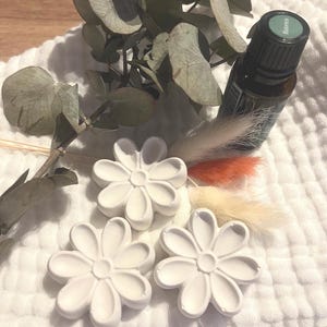 Könnte beinhalten: Drei weiße, margeritenförmige Aromatherapie-Diffusoren auf einem weißen, strukturierten Tuch, mit einer Flasche ätherischen Öls und getrockneten Eukalyptusblättern und dekorativen Gräsern. Die ätherische Ölflasche hat ein grünes Etikett.