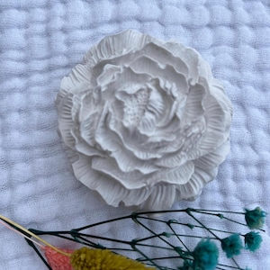 Könnte beinhalten: Ein weißes, strukturiertes, pfingstrosenförmiges Dekorationsobjekt. Die Blume hat detaillierte Blütenblätter und liegt auf einem weißen, zerknitterten Stoff. Getrocknete Blumen in Gelb, Rosa und Blaugrün befinden sich im Vordergrund.