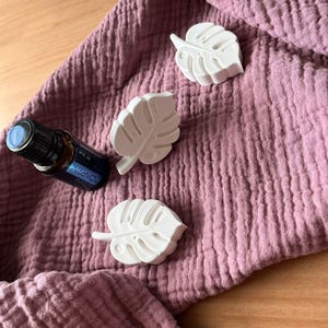 Könnte beinhalten: Drei weiße, blattförmige Dekorationsgegenstände und eine kleine Flasche ätherisches Öl mit blauem Etikett liegen auf einem strukturierten, malvenfarbenen Stoff. Die Objekte sind auf einer Holzoberfläche angeordnet und erzeugen eine natürliche und beruhigende Ästhetik.