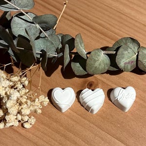Könnte beinhalten: Drei weiße herzförmige Dekorationen mit verschiedenen geprägten Designs, darunter eines mit dem Wort "Love". Die Herzen liegen auf einer Holzoberfläche, begleitet von Zweigen getrockneter Blumen und Eukalyptusblättern.