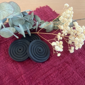 Könnte beinhalten: Drei schwarze Untersetzer aus Gummi mit Spiralmuster auf einem strukturierten bordeauxfarbenen Stoff. Getrockneter Eukalyptus und weiße Blüten ergänzen die Komposition. Die Untersetzer sind rund.