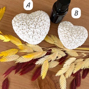 Könnte beinhalten: Zwei weiße herzförmige Objekte mit floralem Muster, neben einer kleinen schwarzen Flasche. Die Herzen liegen auf einer Holzoberfläche mit dekorativen getrockneten Weizenähren in Gelb, Creme und Burgunderrot.