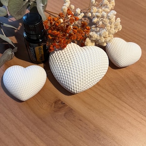 Könnte beinhalten: Ein Stillleben mit drei weißen, herzförmigen Objekten mit strukturierter Oberfläche. Eine kleine Flasche ätherisches Öl und Trockenblumen in Orange und Weiß sind ebenfalls vorhanden. Die Gegenstände sind auf einer Holzoberfläche angeordnet.