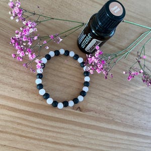 Könnte beinhalten: Ein Armband mit schwarzen und weißen Perlen, eine Flasche ätherisches Öl und rosa Blüten. Die ätherische Ölflasche ist mit "Cedarwood" und "dōTERRA" beschriftet. Das Armband besteht aus schwarzen und weißen runden Perlen.