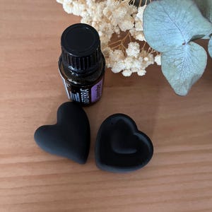 Könnte beinhalten: Eine schwarze ätherische Ölflasche und zwei schwarze herzförmige Objekte sind auf einer Holzoberfläche angeordnet. Die Flasche hat einen schwarzen Deckel und ein Etikett. Getrocknete Blumen und Grünzeug befinden sich im Hintergrund.
