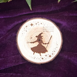 Könnte beinhalten: Ein runder Holzuntersetzer mit einem Hexenmotiv. Der Untersetzer zeigt eine Hexensilhouette mit Hut und Zauberstab, umgeben von Sternen. Der Text "BE THAT WITCH WEAR THAT HAT DO THAT MAGIC" ist ebenfalls enthalten.